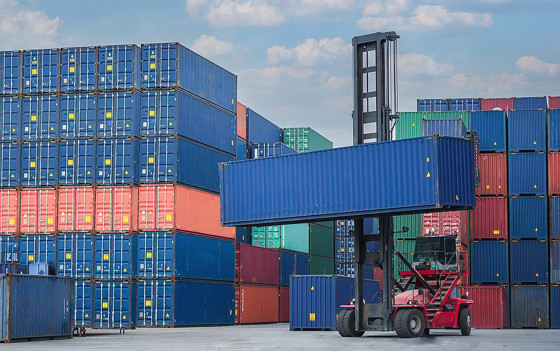 A container handler lifting a blue shipping container in a freight yard stacked with containers