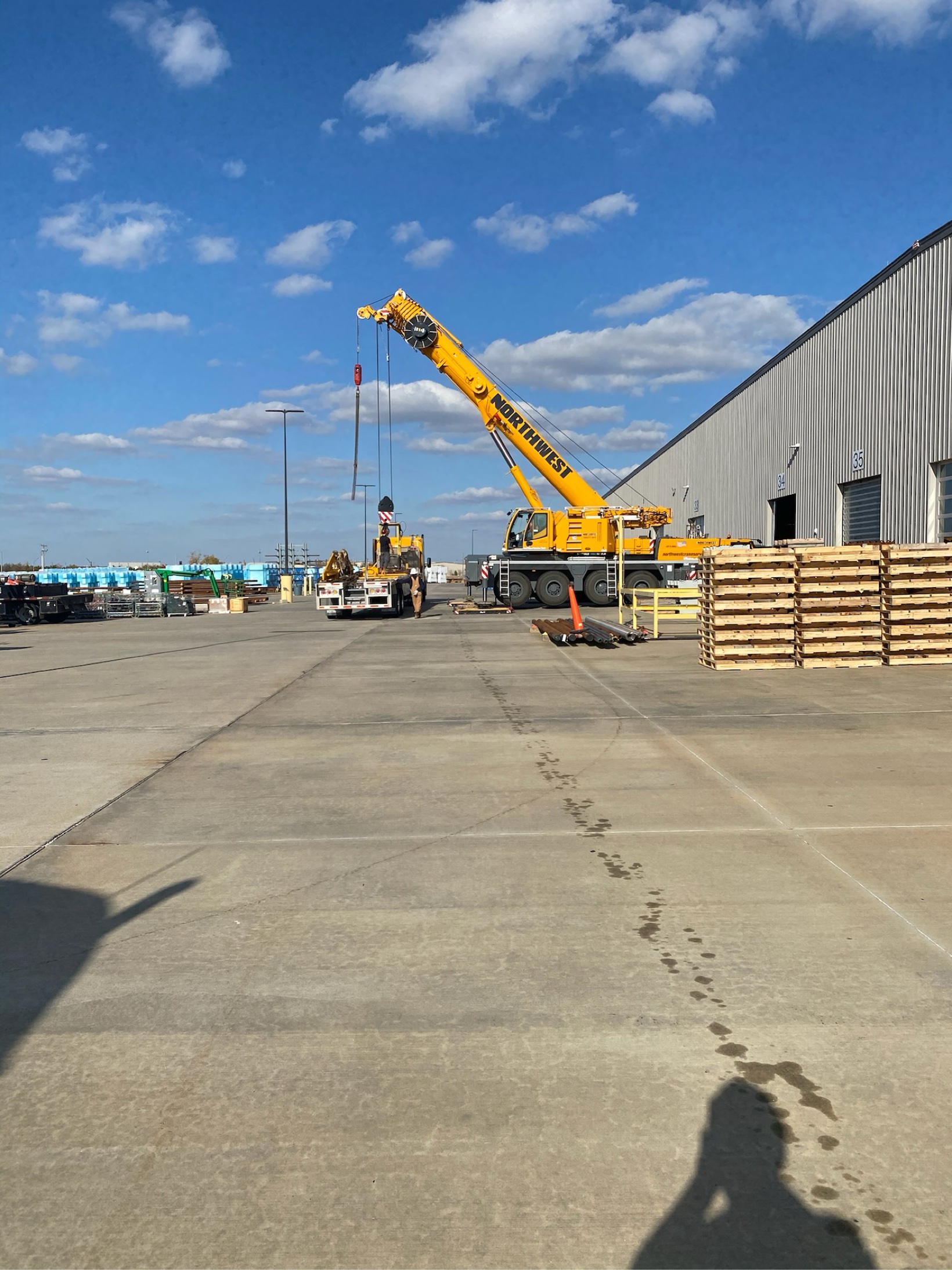Yellow mobile crane lifting a long steel pipe in a warehouse yard